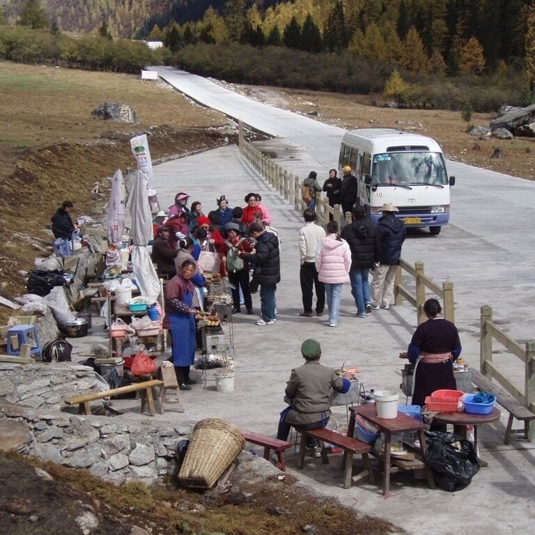 A group of people gathered near a road with a white van, some sitting at tables with goods, and a mountainous landscape in the background.