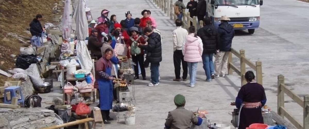 A group of people gathered near a road with a white van, some sitting at tables with goods, and a mountainous landscape in the background.