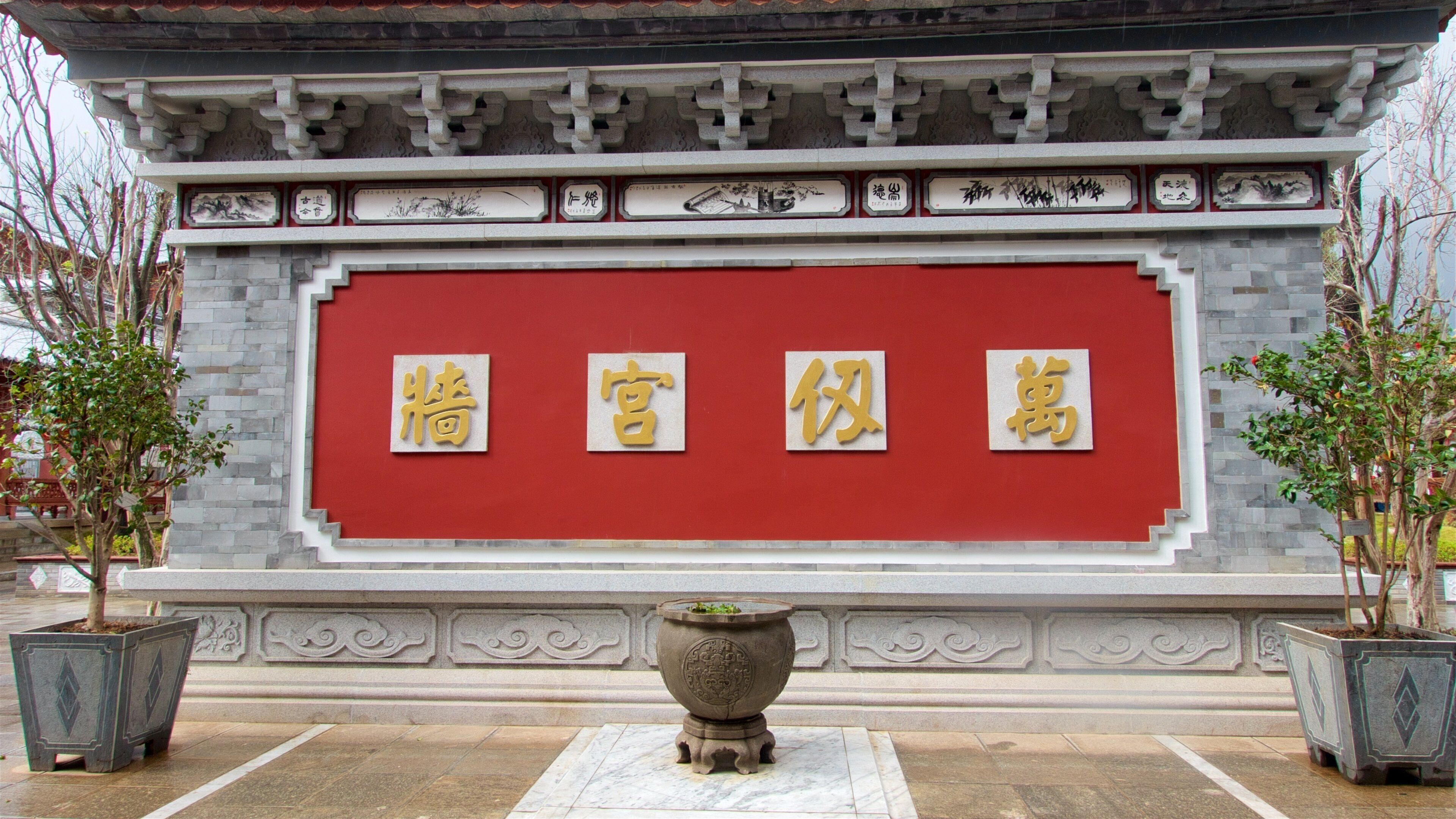 A traditional Chinese building with a red wall and gold Chinese characters. There are two potted plants in front.