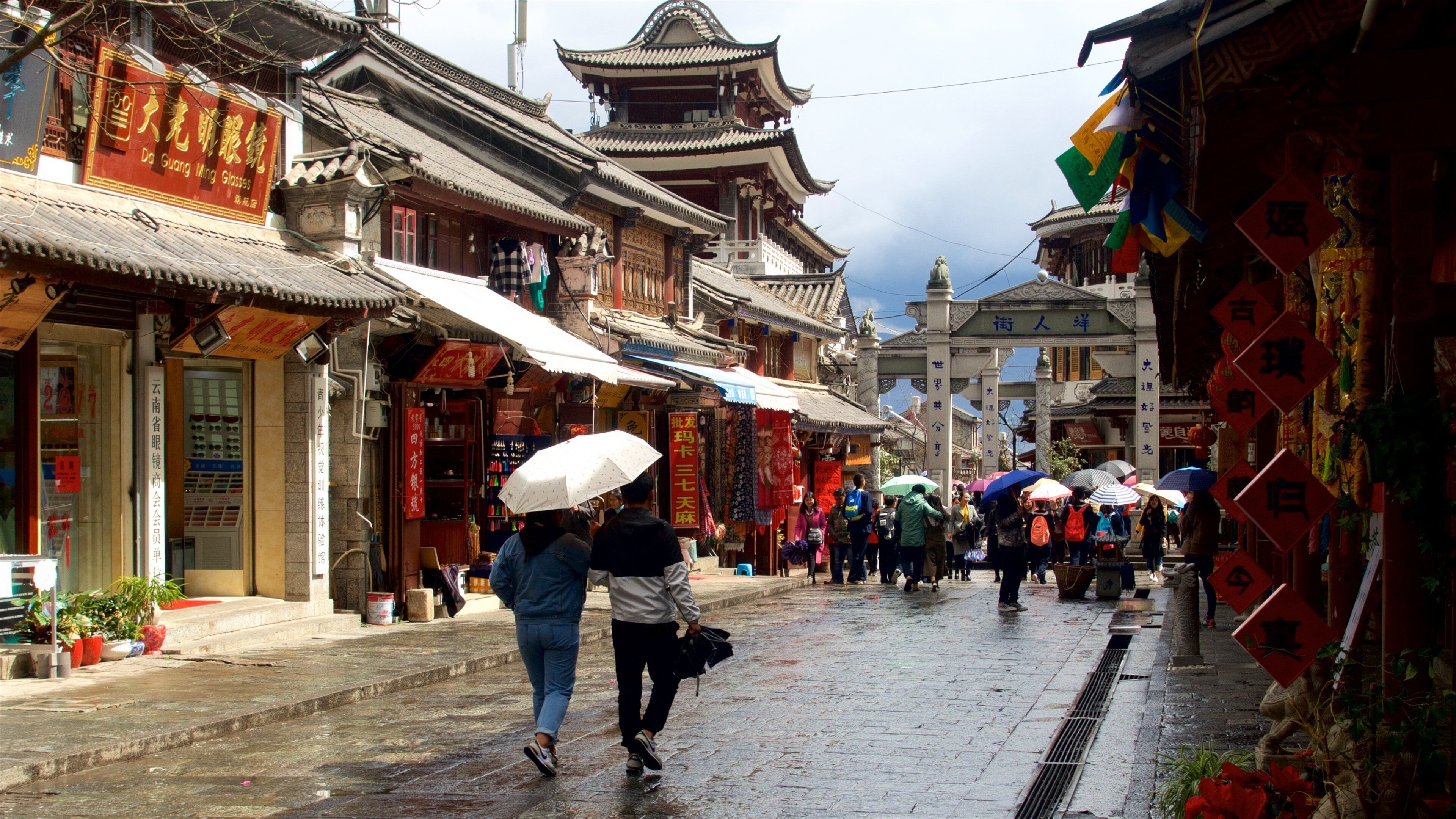 A traditional street with ornate buildings, a large gate, and people walking with umbrellas.