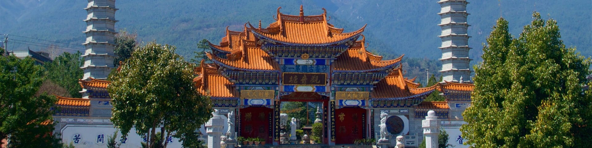 A traditional Chinese gate with two pagoda-style towers, surrounded by lush greenery and mountains in the background.