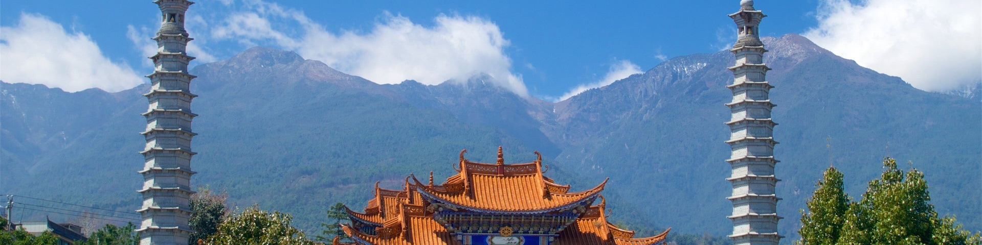 Um portão tradicional chinês com duas torres em estilo pagode, rodeado por vegetação exuberante e montanhas ao fundo.