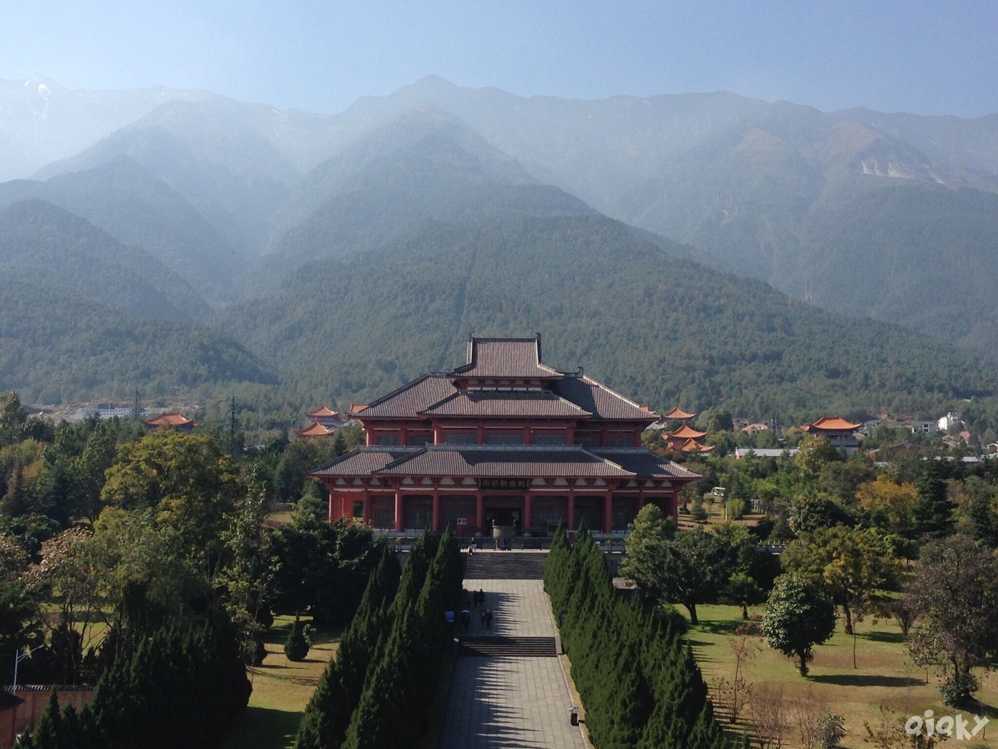 A traditional building with a red roof and white walls, surrounded by a well-maintained garden and trees, with mountains in the background.