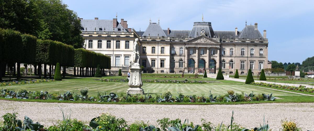 A grand historic building with a symmetrical garden and a statue in the foreground.