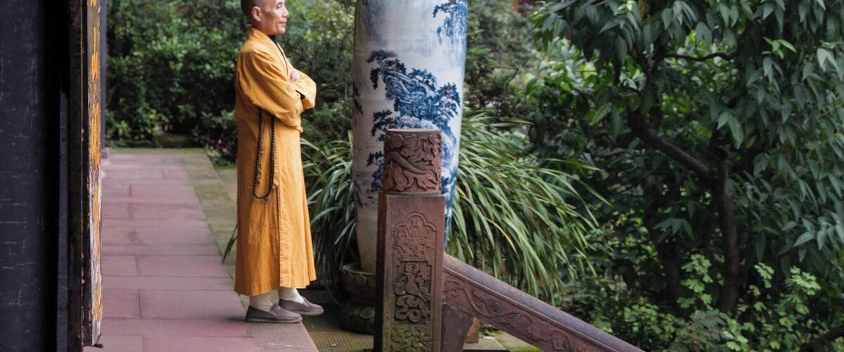 A monk in traditional yellow robes standing on a pathway next to a large blue and white vase.