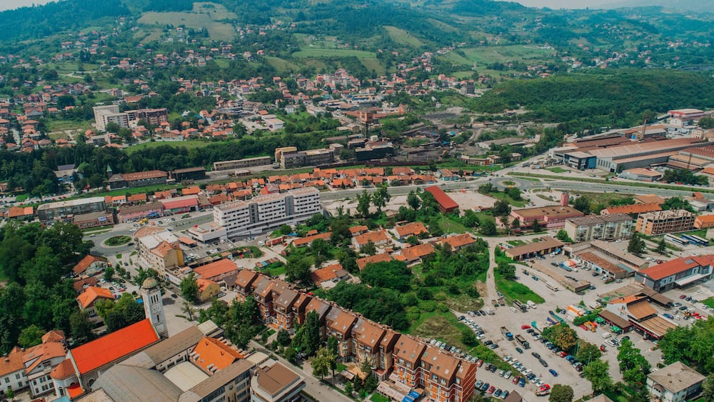 An aerial view of a town with a mix of residential and commercial buildings, surrounded by green hills.