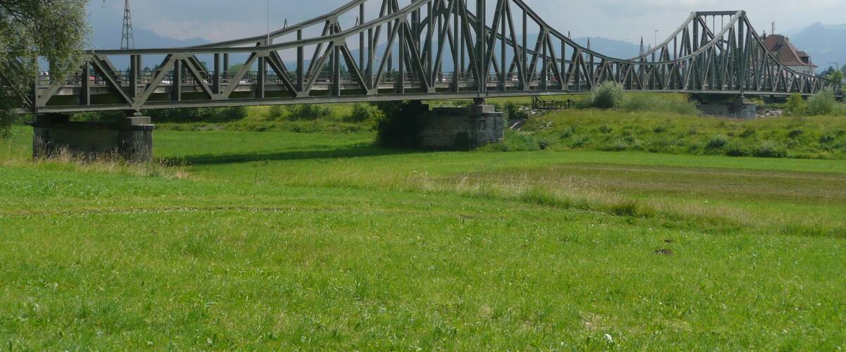 A large steel bridge spanning a river with a mountainous backdrop.
