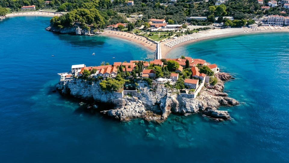 A coastal town with a prominent rocky peninsula, clear blue waters, and a beach.