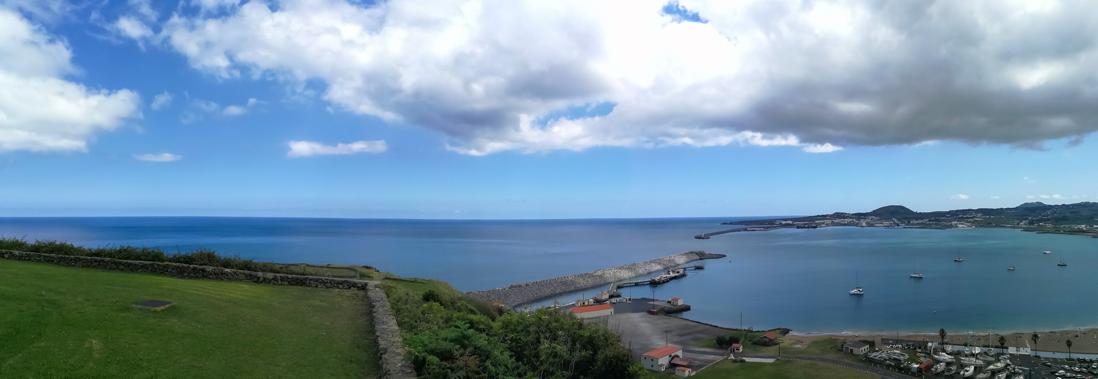 A coastal landscape with a stone wall, grassy area, and a harbor with boats.