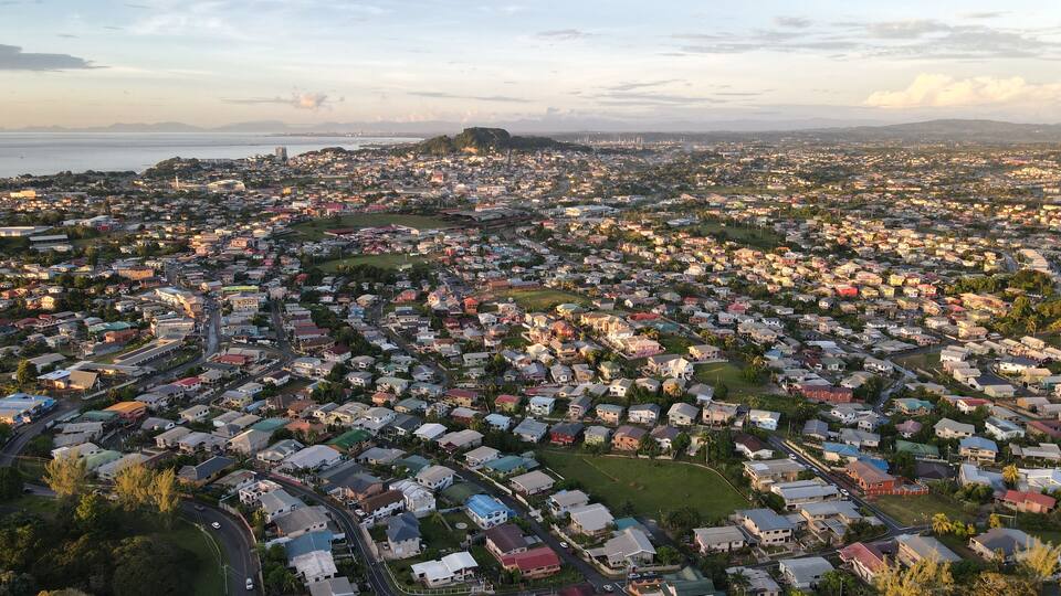 A densely populated urban area with numerous houses and buildings, surrounded by greenery and a coastline in the distance.