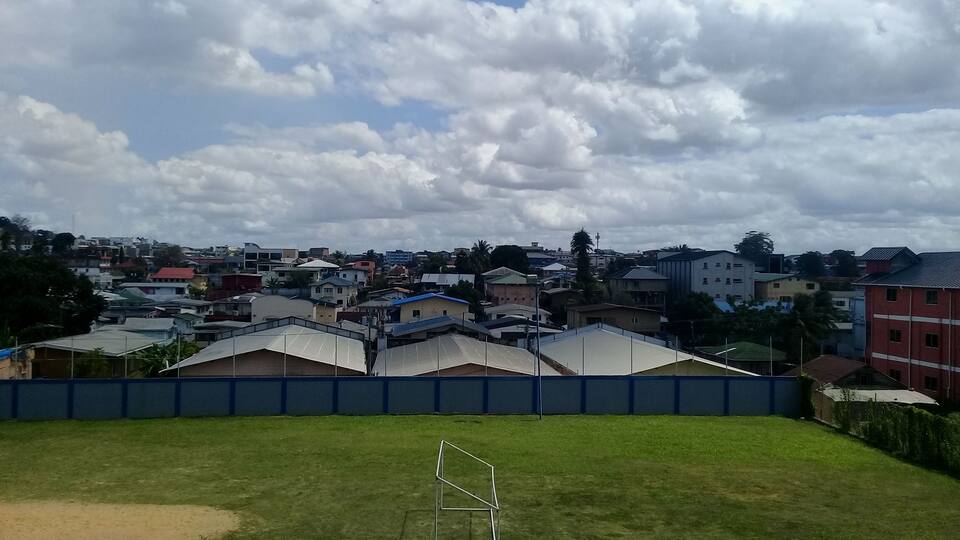 A grassy field with a goalpost, surrounded by a blue fence, and houses in the background under a cloudy sky.