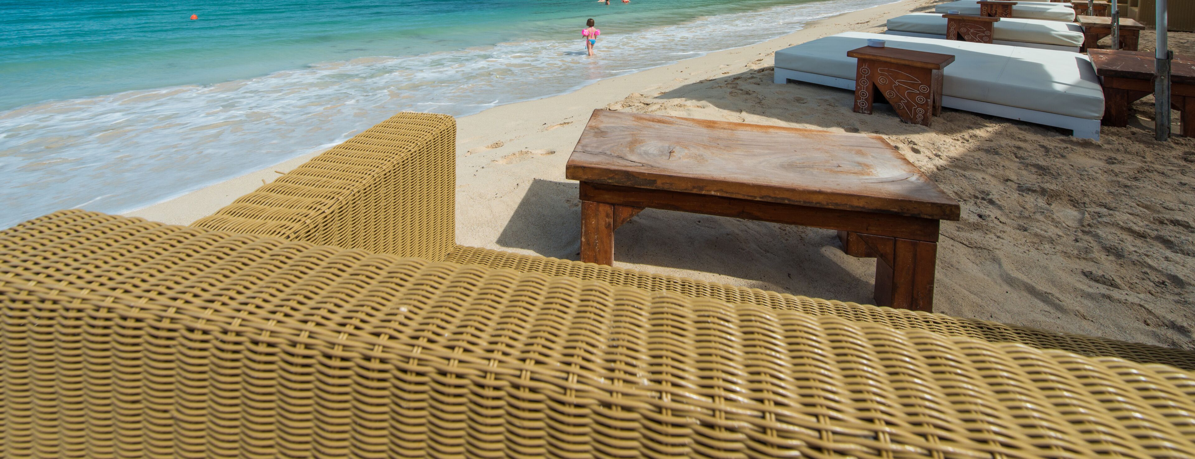 Una playa con sillas reclinables de mimbre, una mesa de madera y asientos acolchados blancos.