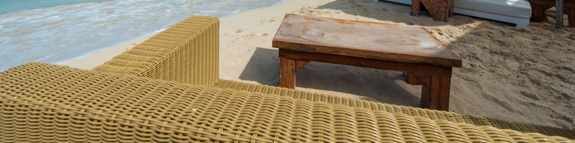 Una playa con sillas reclinables de mimbre, una mesa de madera y asientos acolchados blancos.