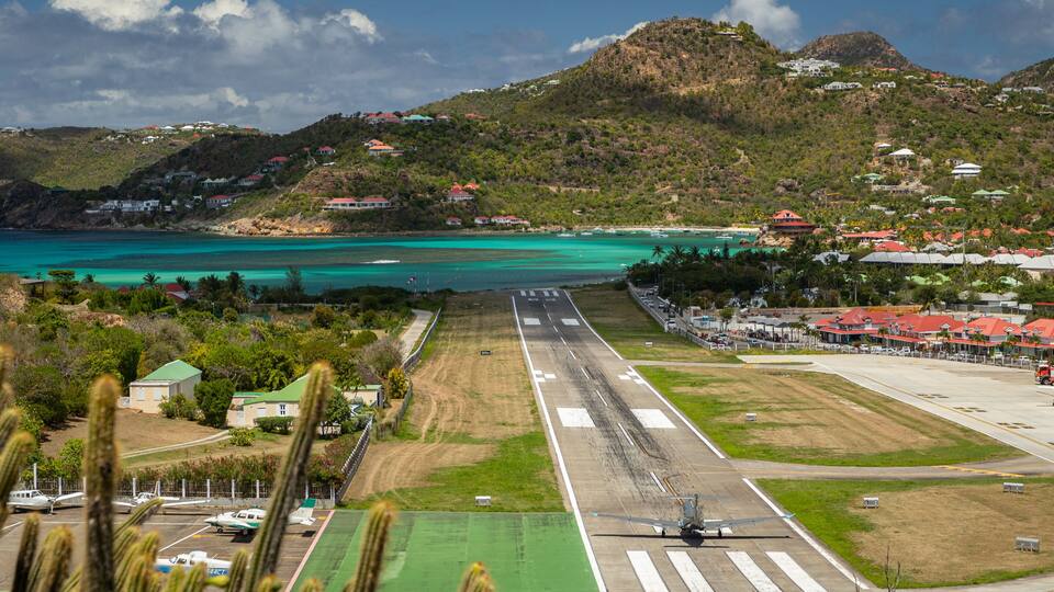 A coastal airport runway with a helicopter approaching for landing, surrounded by lush greenery and houses on a hillside.