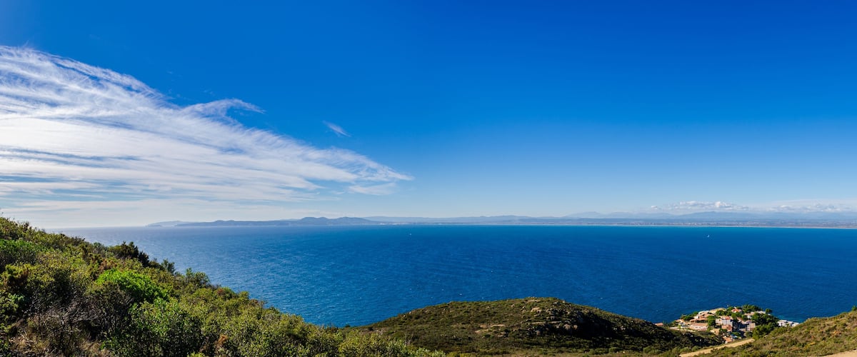Paisagem costeira com mar azul cristalino, uma cordilheira ao longe e um pequeno povoado à direita.