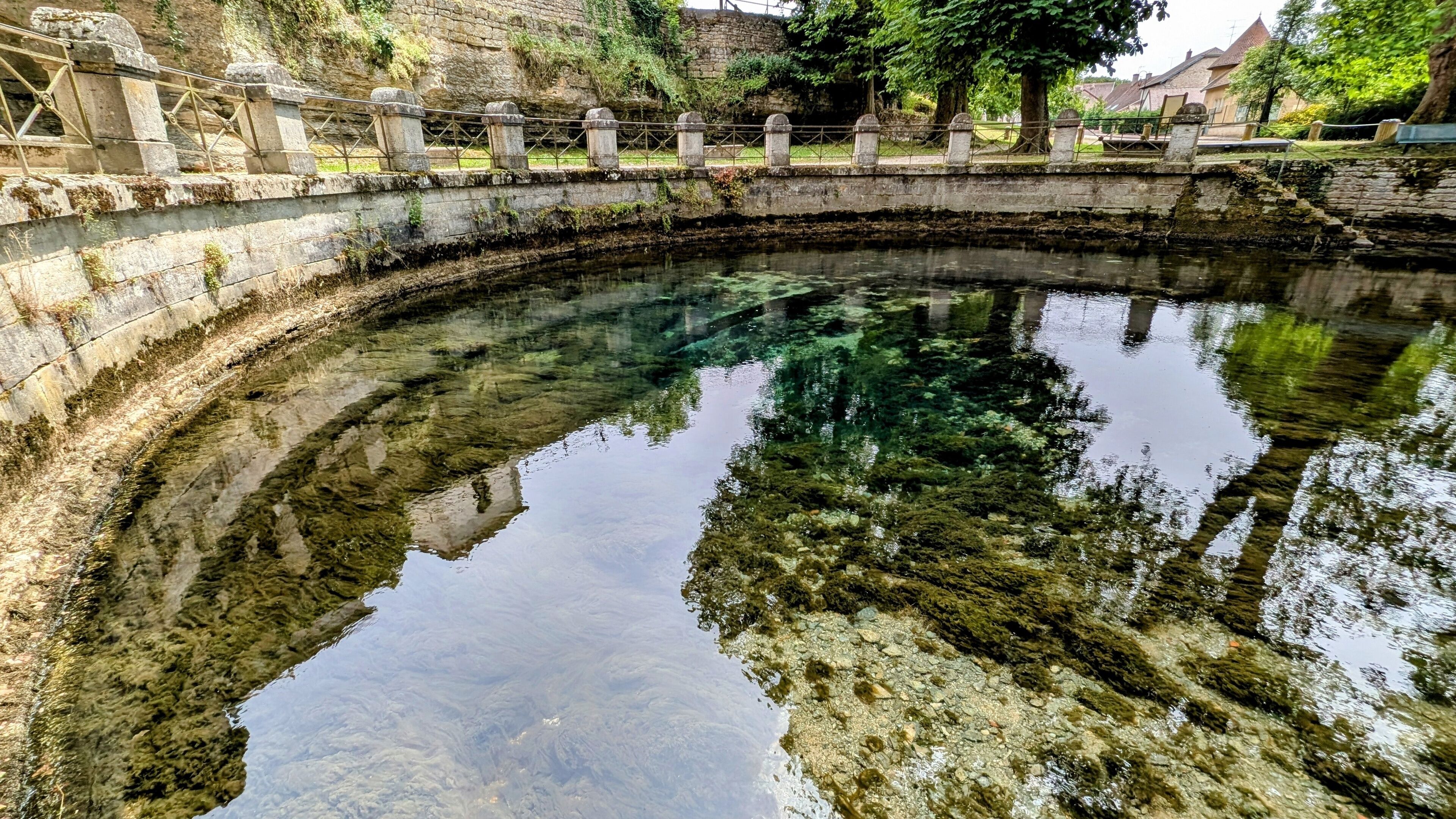 A clear, calm water body with a stone bridge and surrounding greenery.