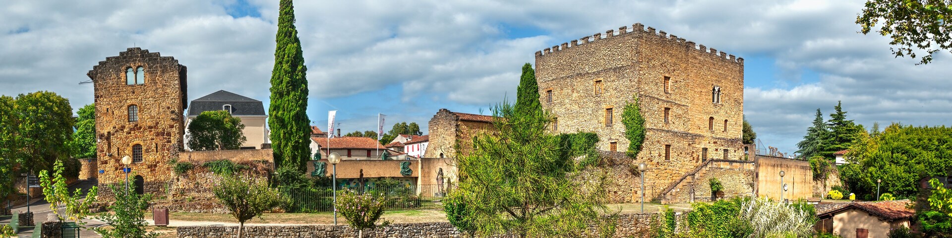 Un castillo de piedra histórico con una torre alta y un gran edificio cuadrado, rodeado de vegetación y un muro de piedra.