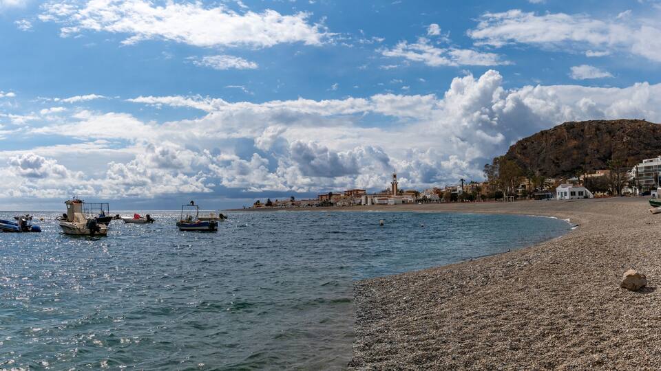 A pebble beach with boats, a rocky hill, and buildings in the background.