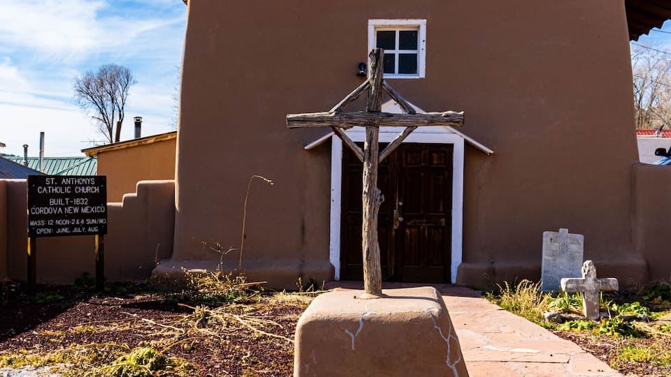 A brown church with a cross on the roof, a wooden cross in front, and a sign in the background.