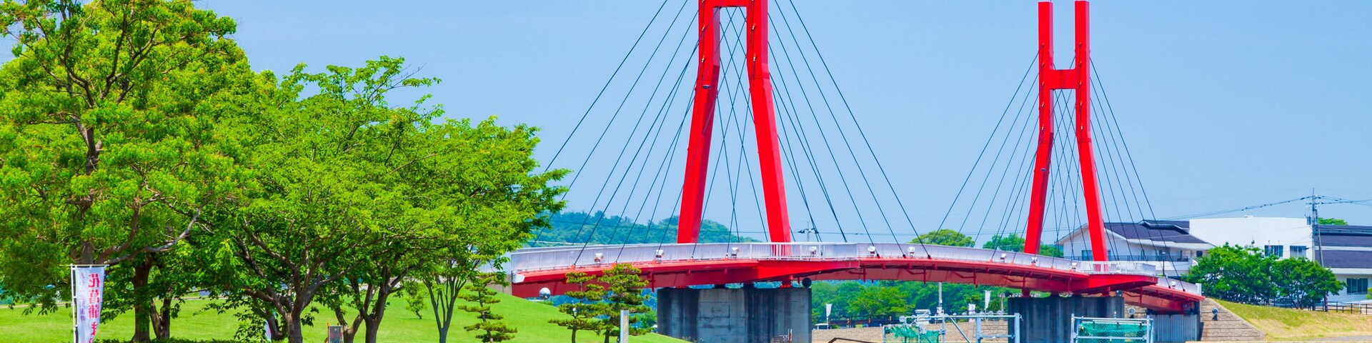 A red suspension bridge over a calm river, surrounded by greenery and a clear sky.