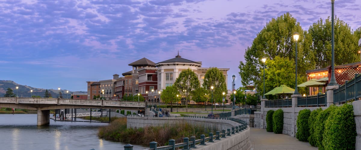 A riverside walkway with a bridge, buildings, and street lamps.