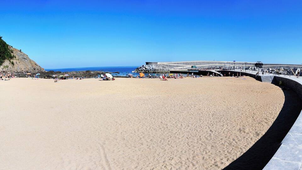 A beach with a rocky cliff, a pier, and people enjoying the seaside.