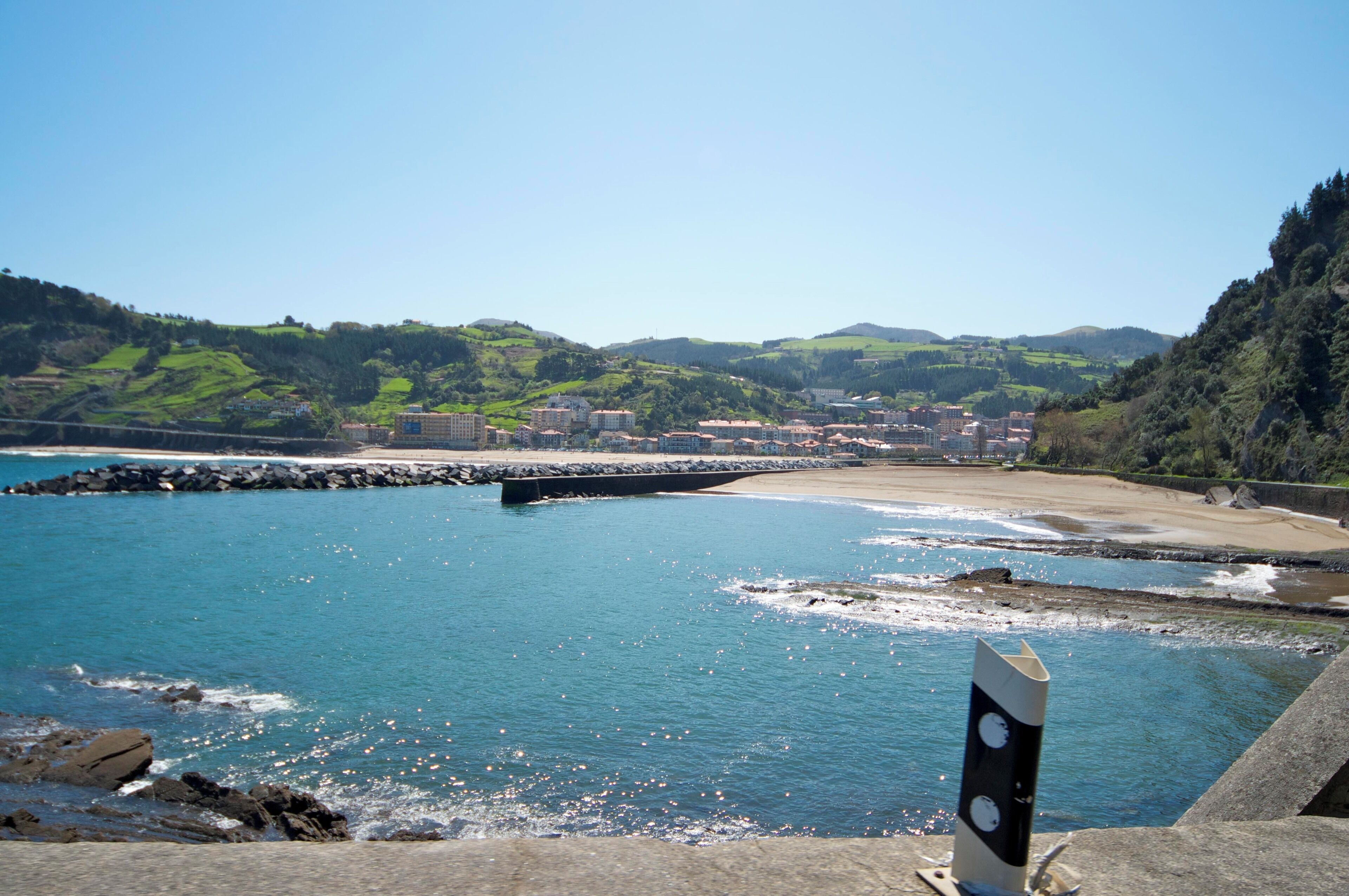 A coastal town with a rocky shoreline, a bridge, and a backdrop of green hills.