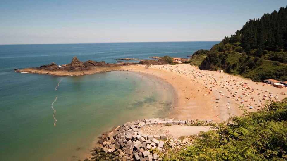 A beach with clear waters, rocky outcrops, and dense forested hills.