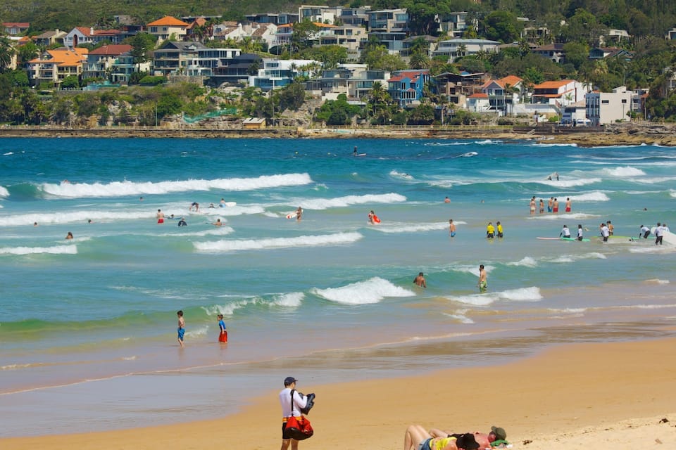 A beach with waves, people, and houses in the background.