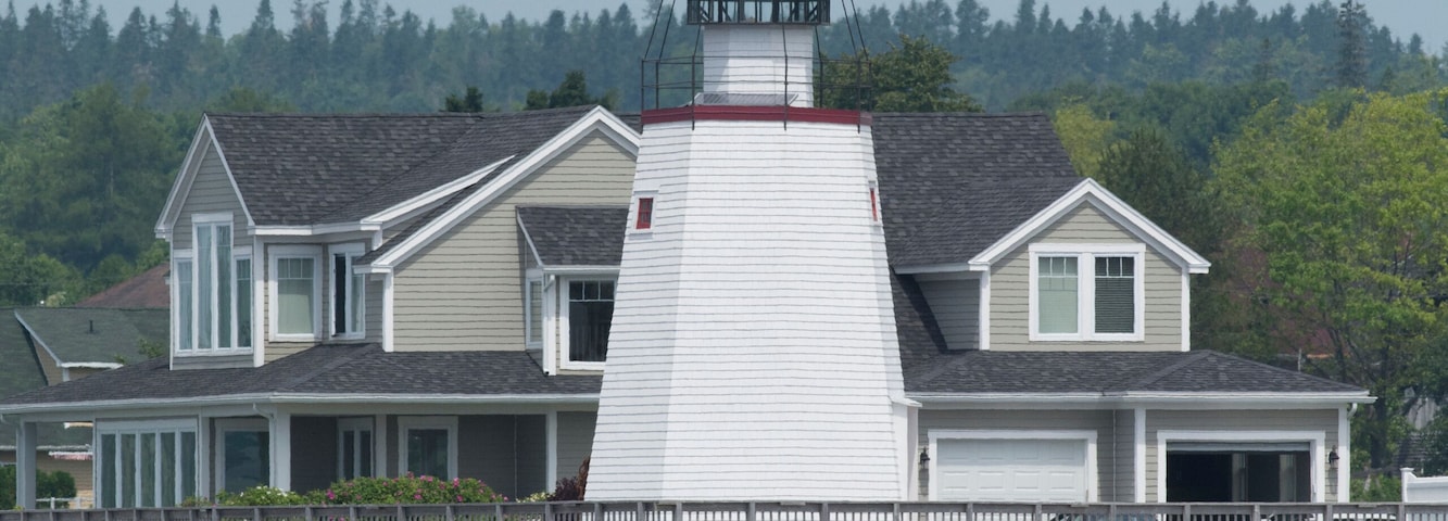 Un faro con la parte superior roja, una casa blanca con techo gris y una costa rocosa.