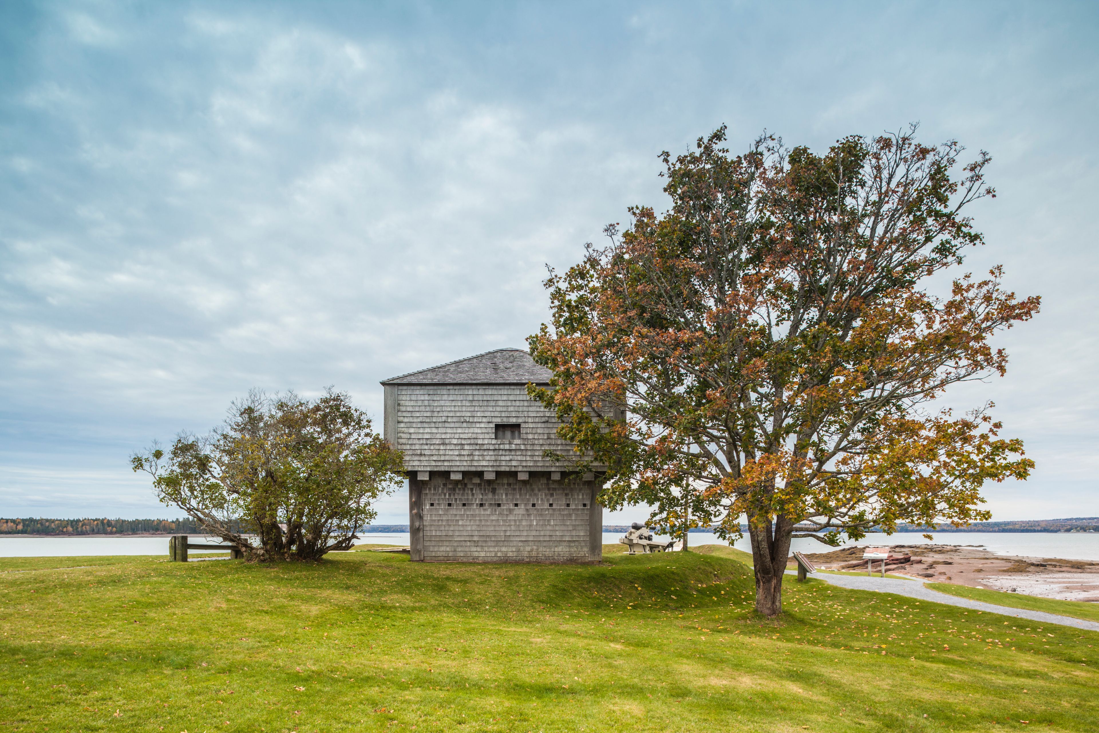 A small wooden structure with a sloped roof, surrounded by grass and trees with autumn leaves, near a body of water.