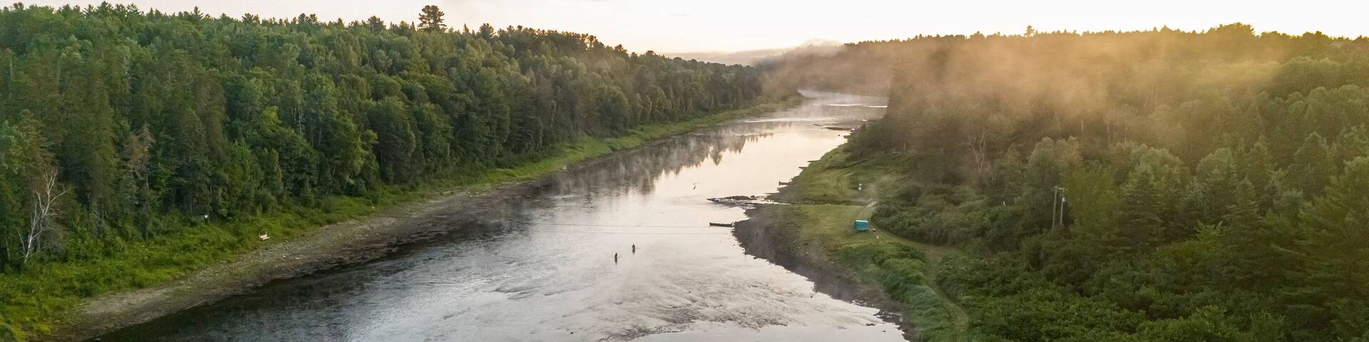A river surrounded by dense forest, with a misty atmosphere and a sunrise or sunset.