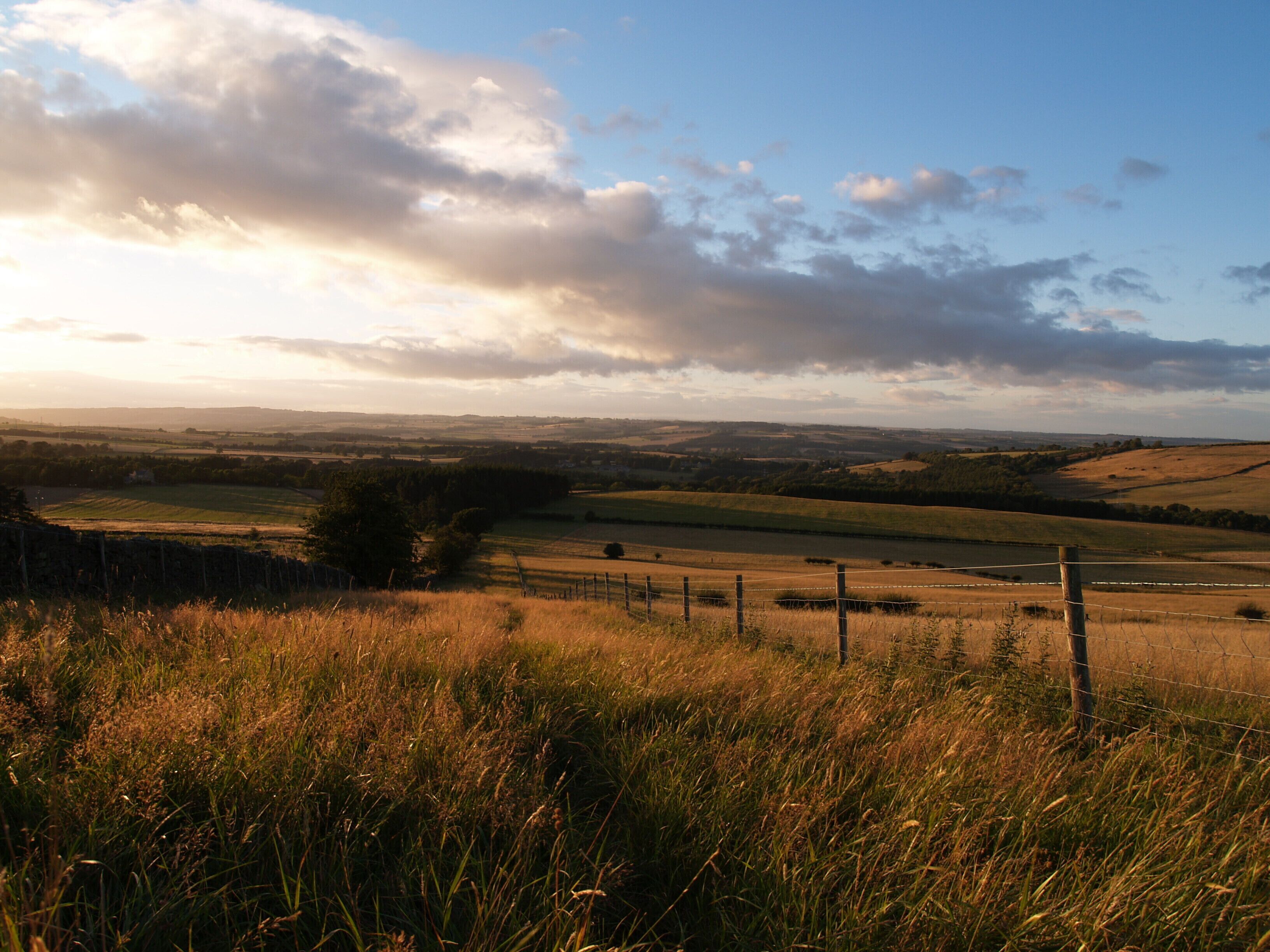 A rural landscape with rolling hills, a river, and a fence in the foreground.