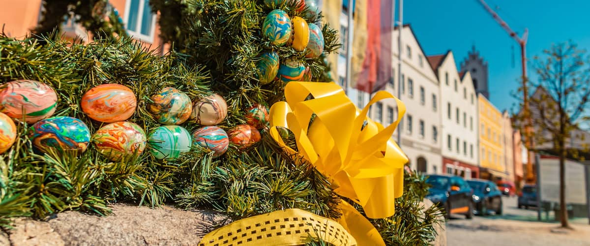 A decorated Christmas tree with colorful ornaments and a yellow bow, set against a backdrop of European buildings and a clear blue sky.