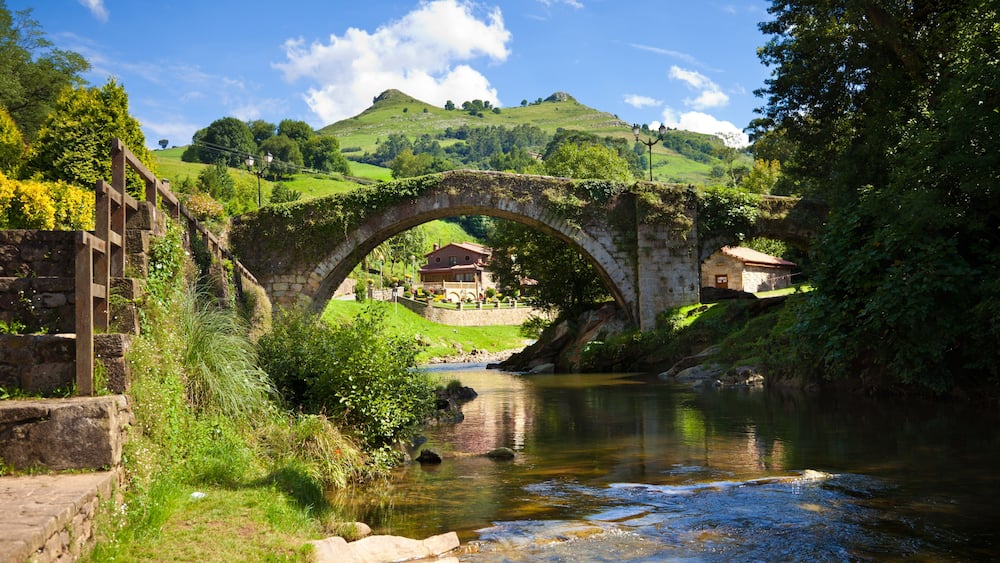 A stone bridge over a river, surrounded by greenery and hills.