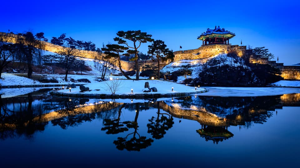 A snow-covered landscape with a traditional pagoda and stone walls at dusk.