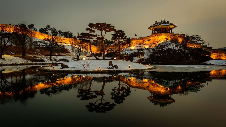 A snow-covered landscape with a traditional pagoda, a stone wall, and a calm body of water reflecting the structures.