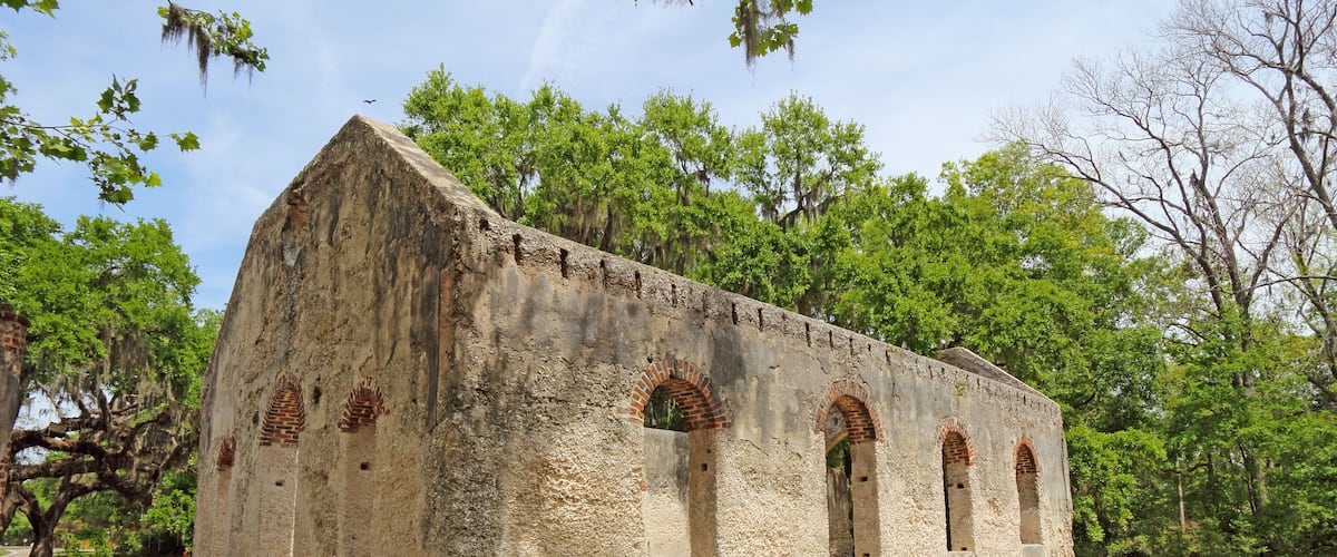An old stone building with arched windows and a brick roof, surrounded by trees and grass.