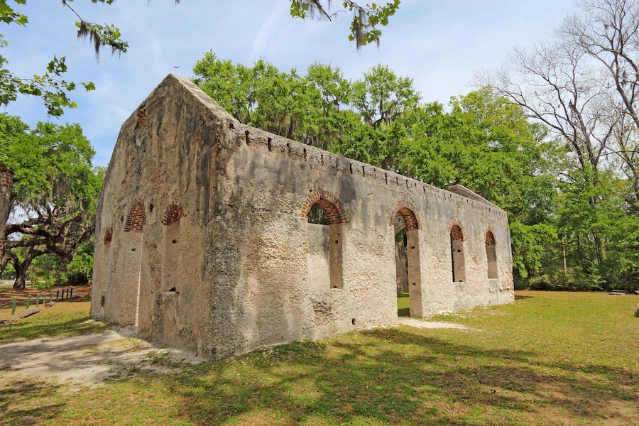 Un antiguo edificio de piedra con ventanas de arco y techo de ladrillo, rodeado de árboles y césped.