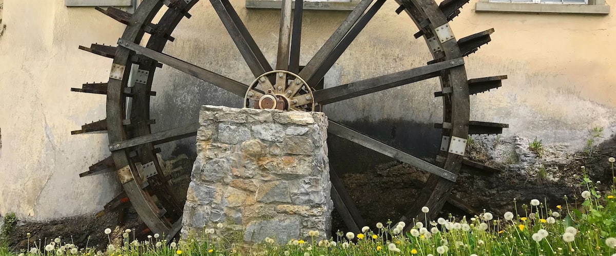 A large water wheel with a stone base, set against a building with windows.