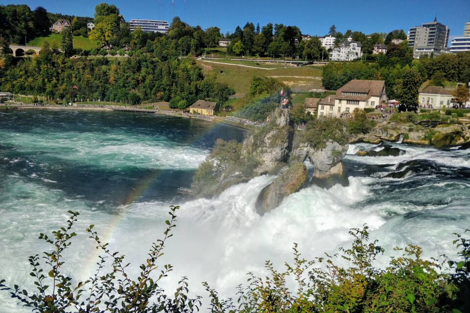 A river with a waterfall, surrounded by greenery and buildings.