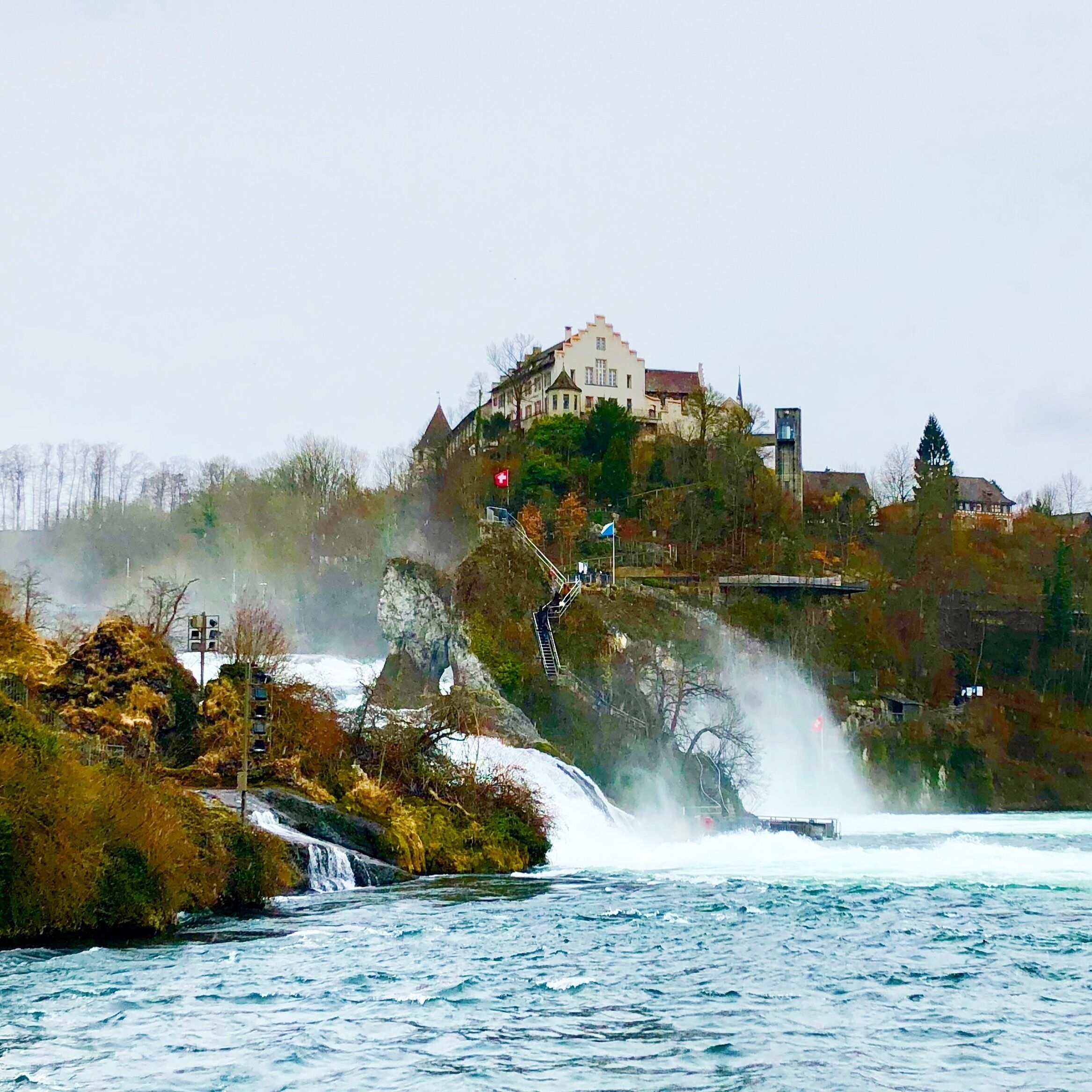 A waterfall cascading down a rocky cliff into a river, with a historic building on a hill in the background.
