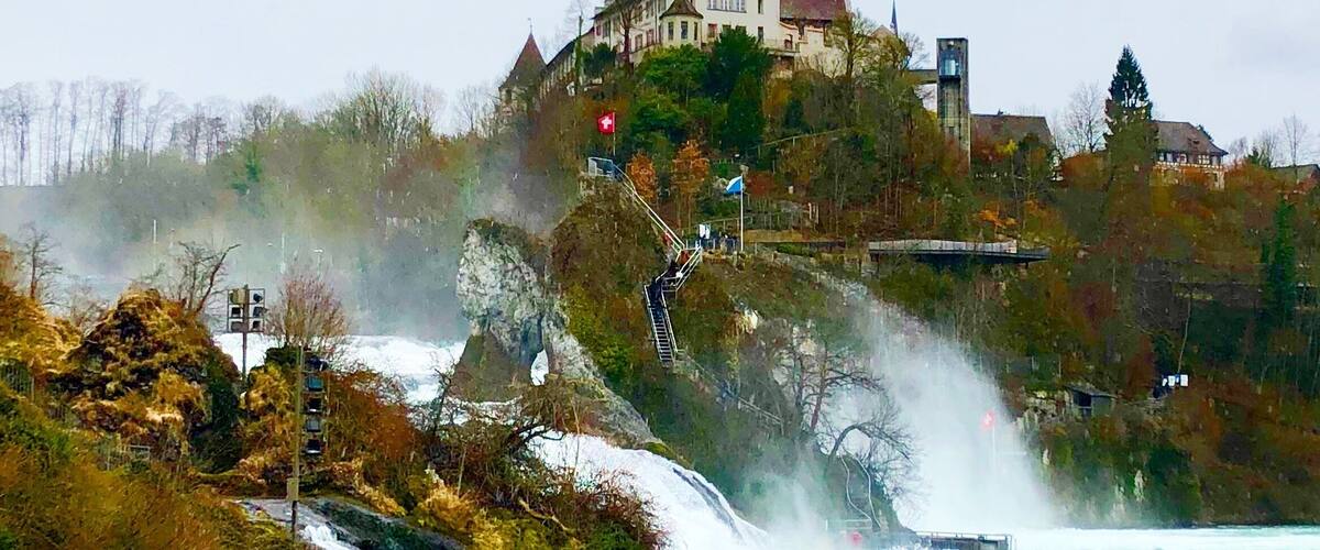 A waterfall cascading down a rocky cliff into a river, with a historic building on a hill in the background.