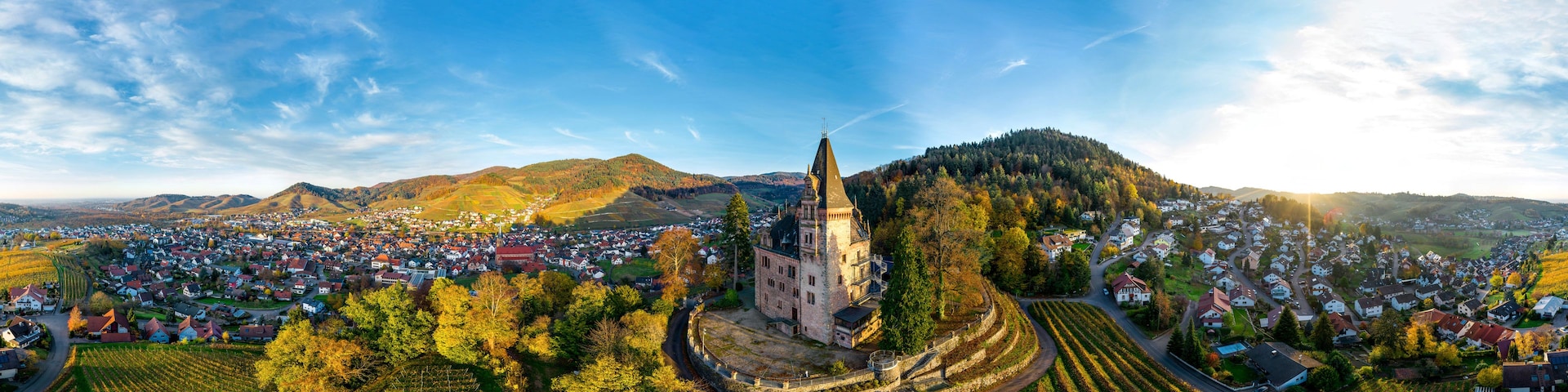 Una vista panorámica de un pueblo con un castillo destacado, viñedos en terrazas y colinas ondulantes al fondo.