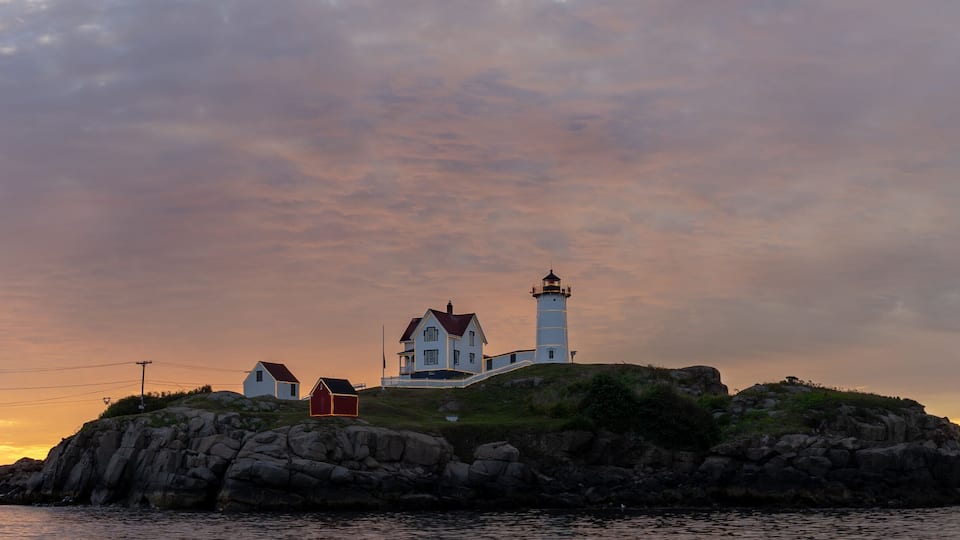 A coastal scene with a lighthouse on a rocky outcrop, surrounded by houses and a sunset sky.