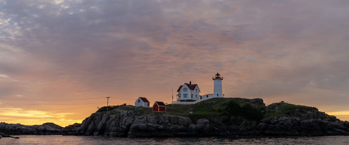 A coastal scene with a lighthouse on a rocky outcrop, surrounded by houses and a sunset sky.