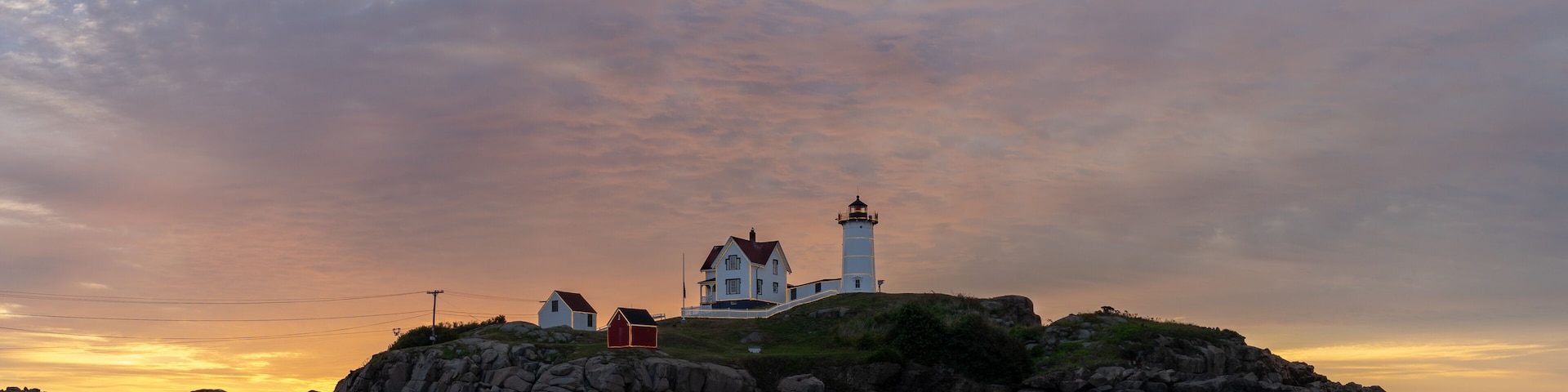 A coastal scene with a lighthouse on a rocky outcrop, surrounded by houses and a sunset sky.