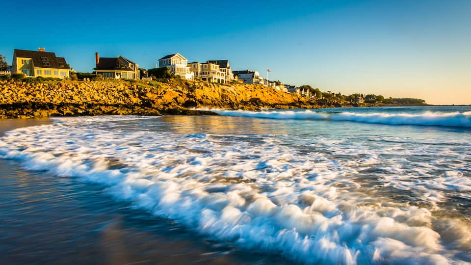 A coastal landscape with waves, a rocky shoreline, and houses on the cliff.