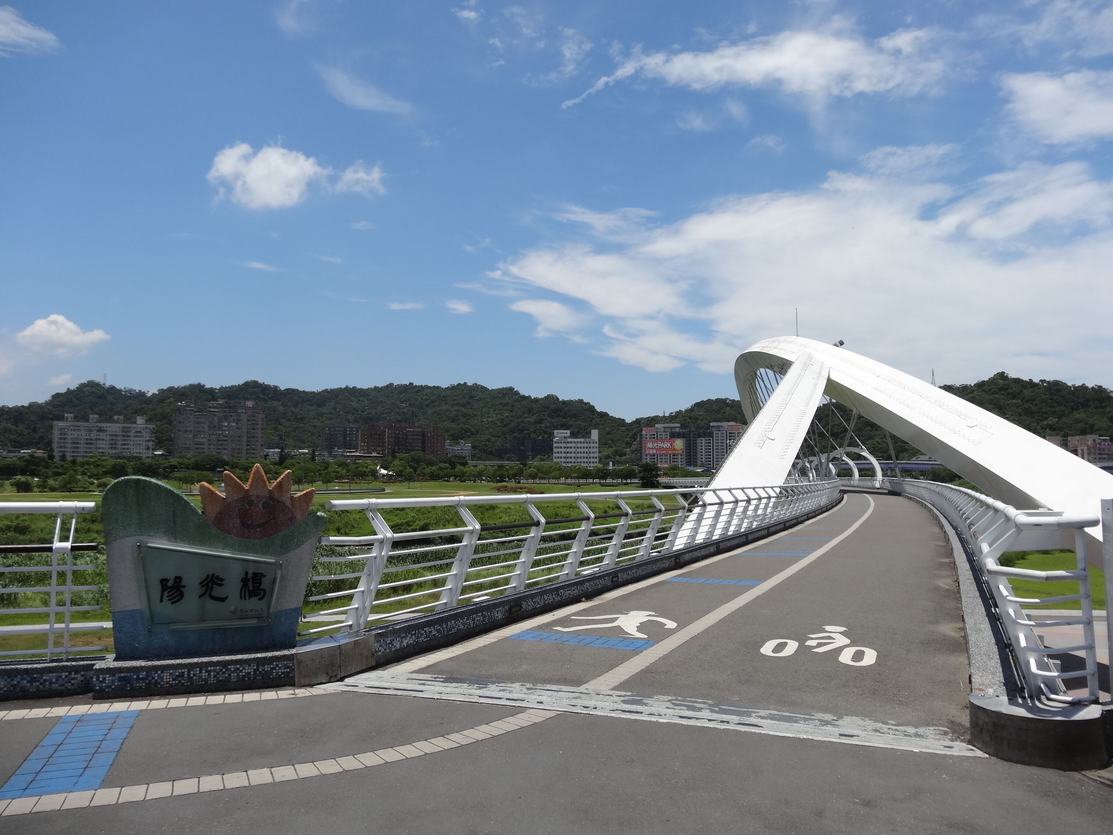 Uma ponte moderna com design de arco branco, ciclofaixa e área verde com um parque e uma colina ao fundo.