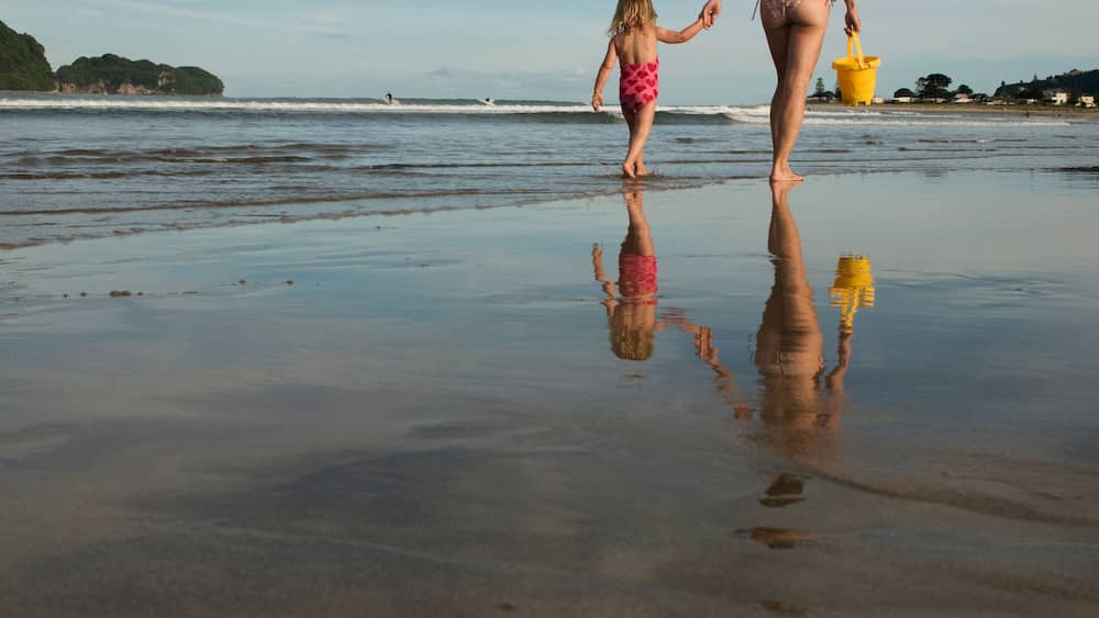 Une plage où un enfant et un adulte se tiennent la main, un seau jaune et un reflet sur le sable mouillé.